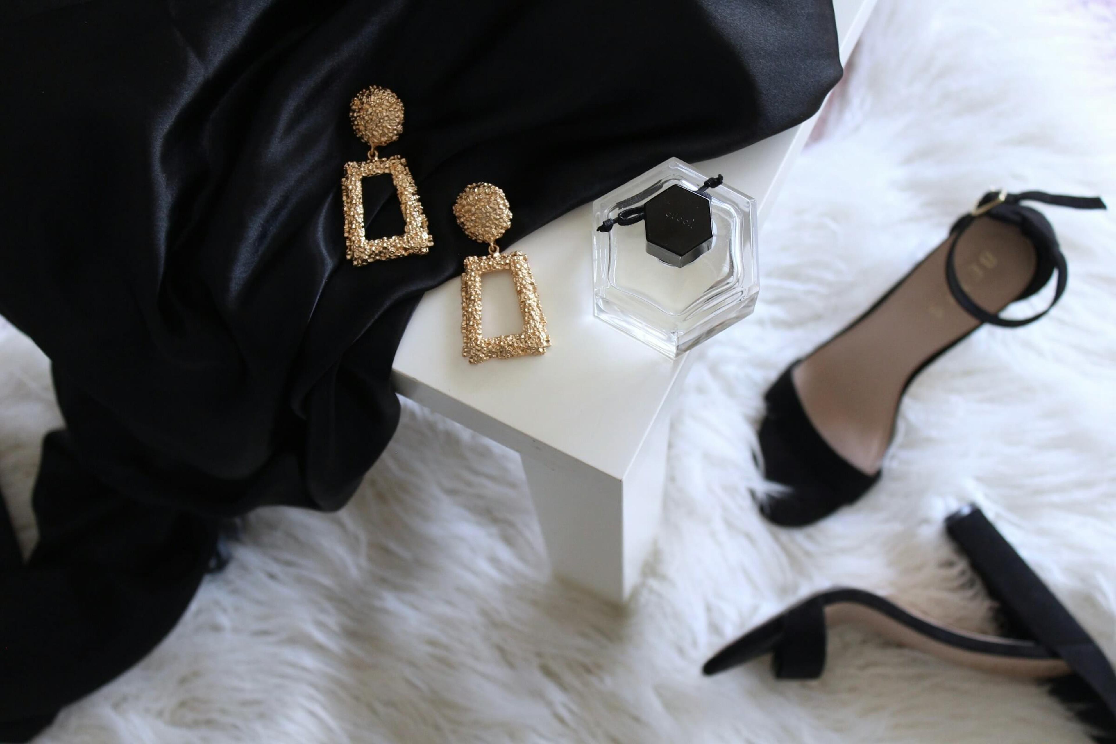 Woman's gold earrings, black dress, black high heels and perfume on white furry carpet